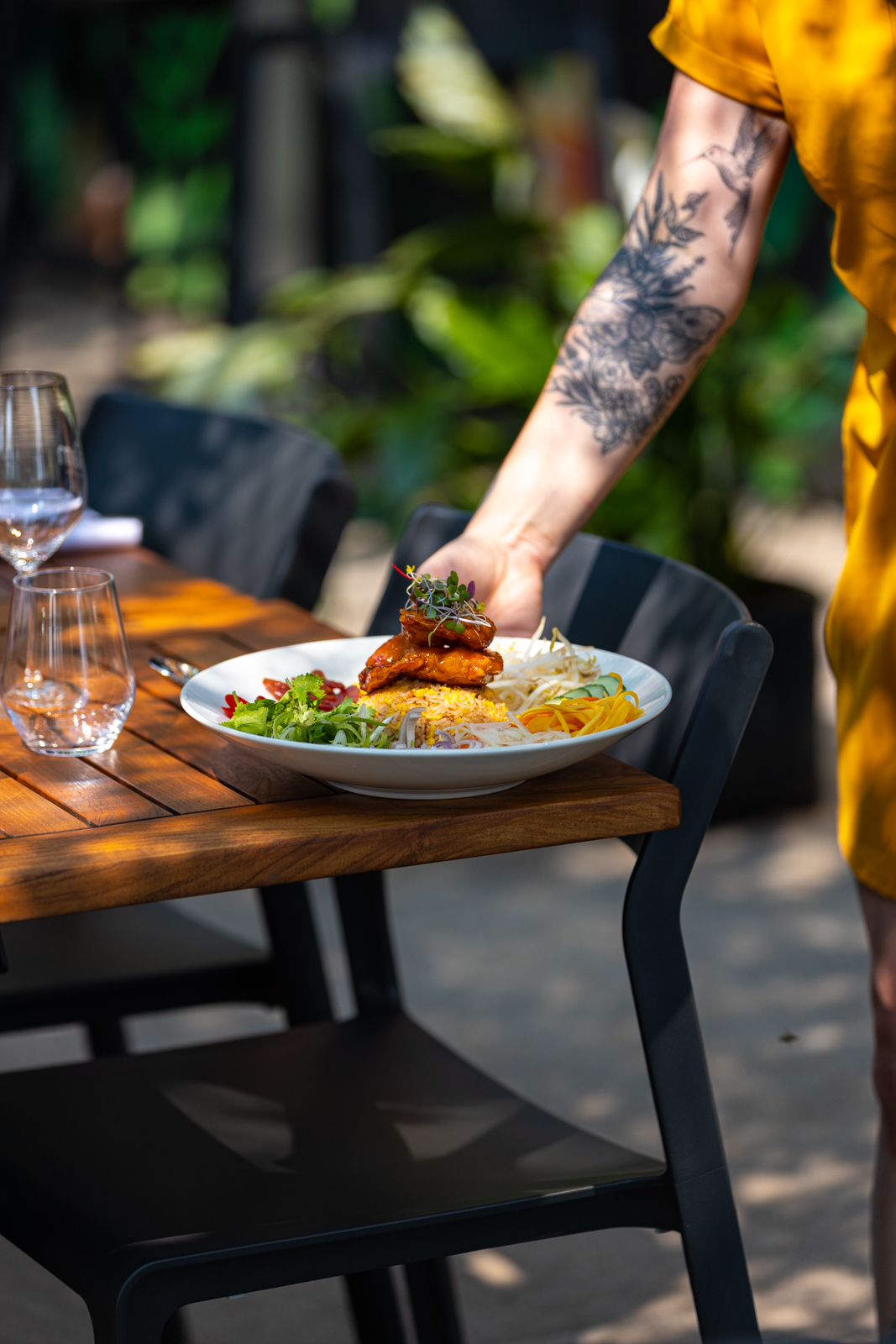 Une dame avec avant-bras tatoué dépose une assiette sur une table en terrasse